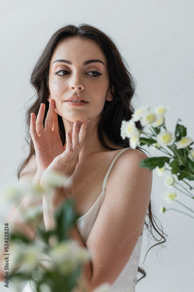 Young woman touching face and looking at blurred flowers isolated on grey. Young woman touching face and looking at blurred flowers isolated on grey.