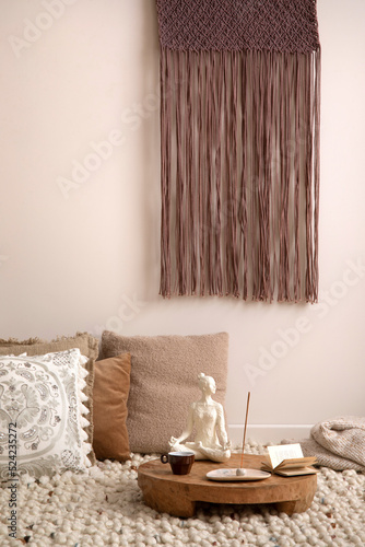 Fototapeta Naklejka Na Ścianę i Meble -  Boho and cozy interior of meditation space with a lot of pillows, plaid, hanging macrame, wooden tray with accessories, decoration, carpet and personal stuff. Warm and minimalist home decor. Template.
