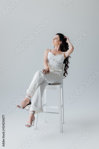 Foto Full length of young model in white clothes and heels sitting on chair on grey background