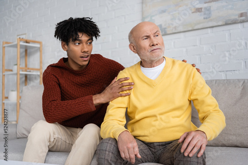 African american grandson hugging disappointed grandpa on couch at home.