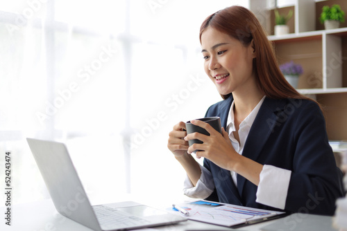 Happy beautiful Asian businesswoman working with laptop in home office and drinking coffee in the morning.