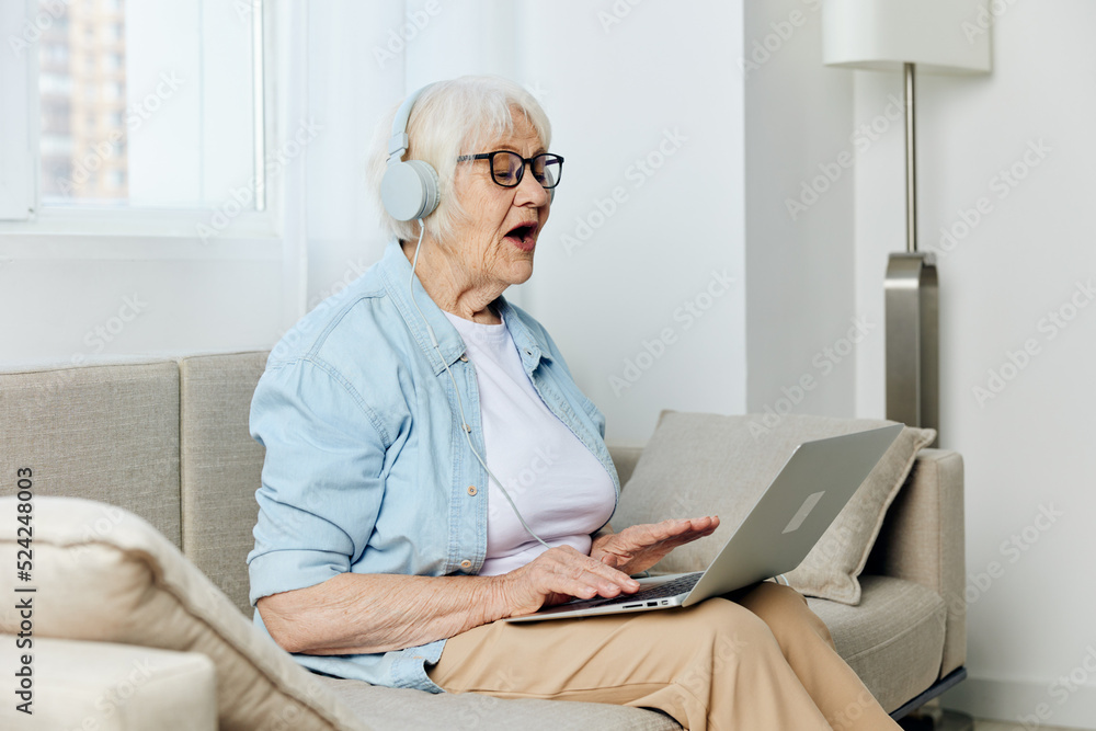 a pleasant, happy elderly woman is sitting on a beige sofa and holding a laptop on her lap, communicating via video using headphones