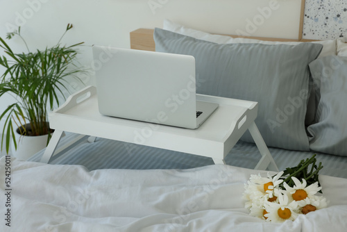 White tray table with laptop and bouquet of beautiful daisies on bed indoors