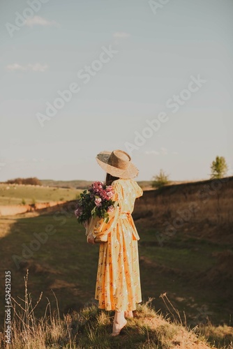 Beautiful girl in a yellow dress in a straw hat and with a bouquet of lilacs in a straw bag stands in the sun on the hill. Back view. 