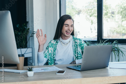 Cheerful queer designer having video call on laptop in creative agency.