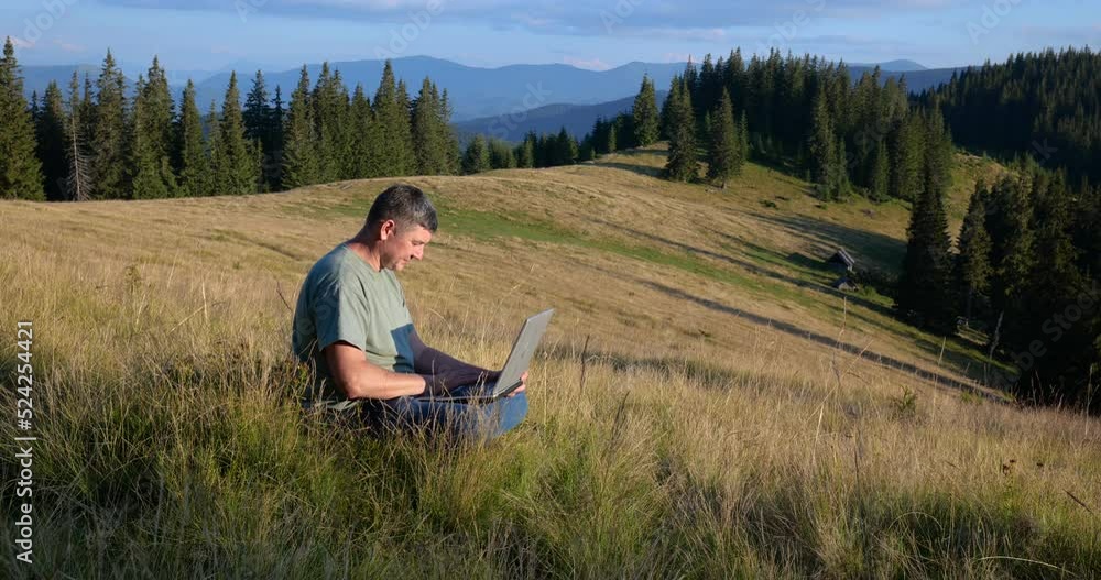 A man sits on a beautiful meadow in the mountains, works on a laptop A man sits on a beautiful meadow in the mountains, works on a laptop