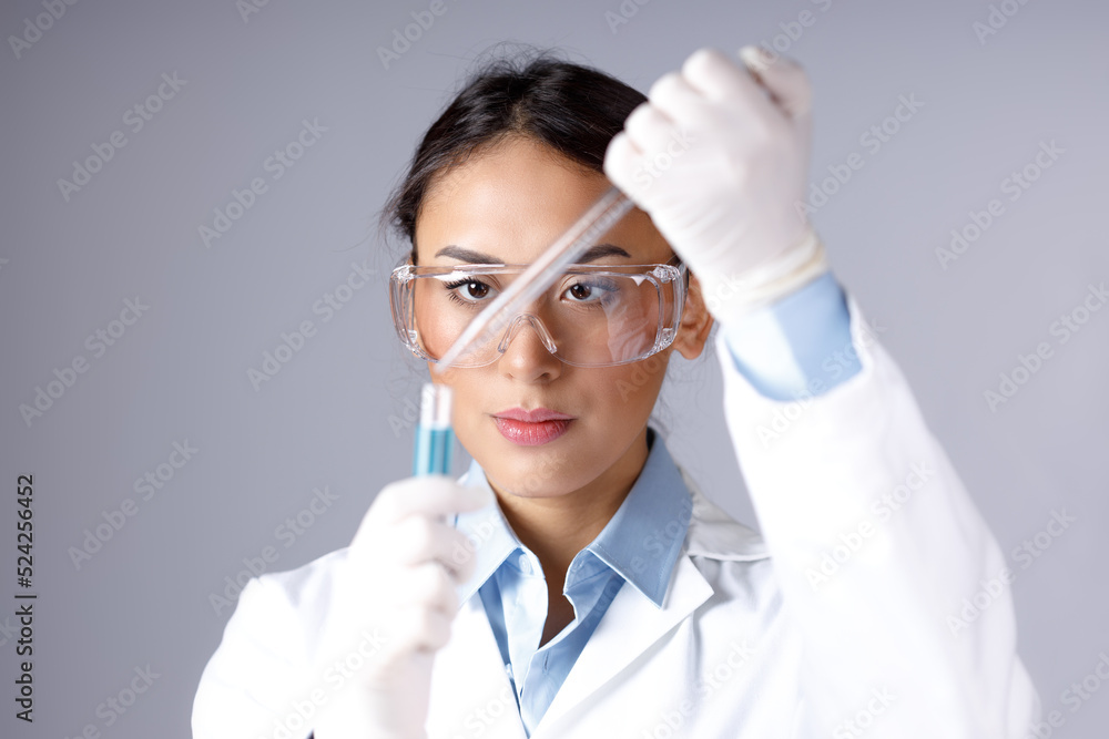 Young female chemist in lab coat, gloves and protective eyeglass dropping a blue substance into a test tube with a pipette. Chemical, pharmaceutical or medical research concept. Young female chemist in lab coat, gloves and protective eyeglass dropping a blue substance into a test tube with a pipette. Chemical, pharmaceutical or medical research concept.
