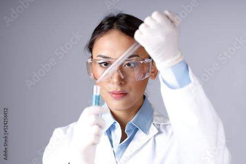 Young female chemist in lab coat, gloves and protective eyeglass dropping a blue substance into a test tube with a pipette. Chemical, pharmaceutical or medical research concept.