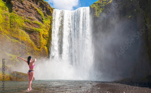 Beautiful girl view from back in bikini raising arms in front of waterfall - Icelandic Landscape concept - View of famous Skogafoss waterfall 