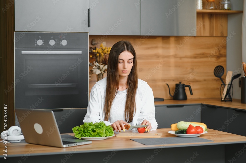 Athletic woman blogger nutritionist prepare a salad with fresh vegetables and conducts a video conference on healthy eating on laptop in the kitchen