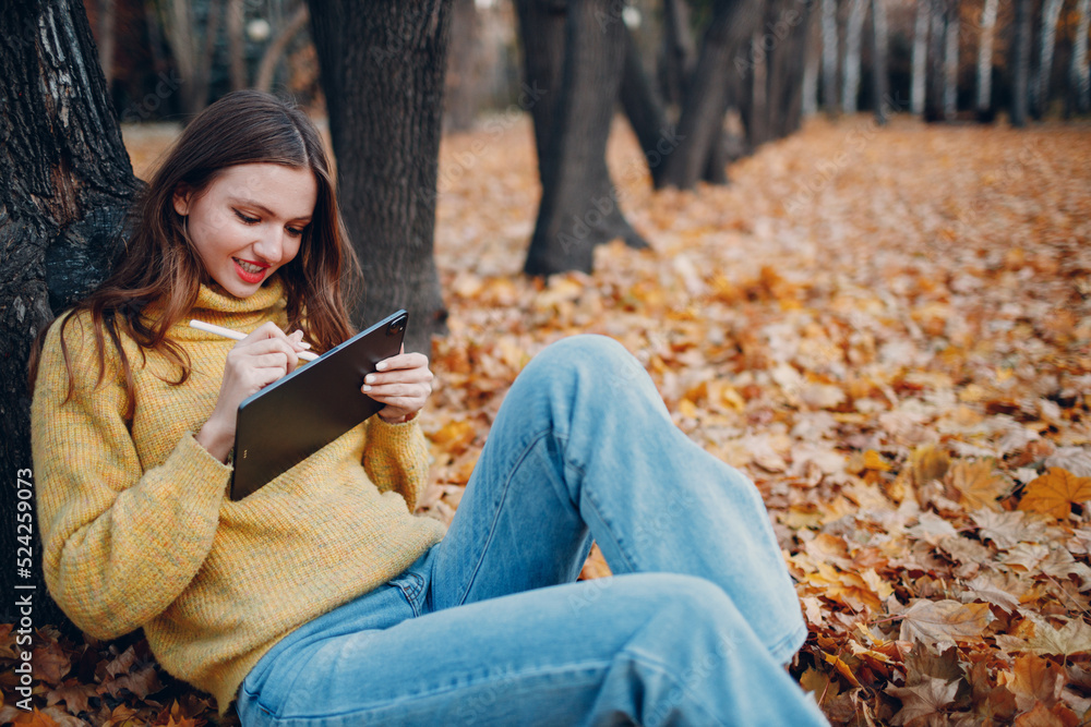 Young woman model sitting with digital tablet and painting with pencil stylus in autumn park with yellow foliage maple leaves. Young woman model sitting with digital tablet and painting with pencil stylus in autumn park with yellow foliage maple leaves.