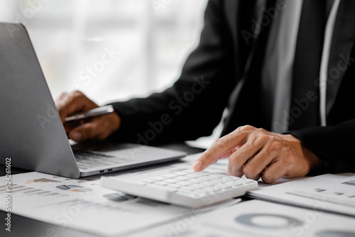Businessman is using a calculator to calculate company financial figures from earnings papers, a businessman sitting in his office where the company financial chart is placed. Banner with copy space.