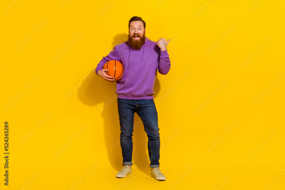 Full length photo of funny impressed guy dressed purple sweater holding ball pointing thumb empty space isolated yellow color background Full length photo of funny impressed guy dressed purple sweater holding ball pointing thumb empty space isolated yellow color background
