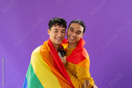 Portrait of happy biracial male couple embracing and holding lgbt flag on purple background