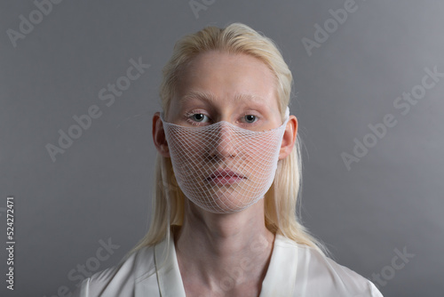 Closeup portrait of white caucasian albino blond woman fashion model wearing quarantine medical face mask coarse mesh net on grey background. Covid-19 coronavirus protection concept.