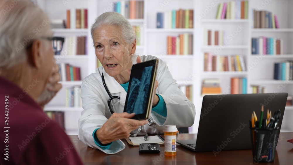 Serious old senior female general practitioner in lab coat holding digital computer tablet, showing laboratory test results to interested elderly 70s male patient at checkup meeting in hospital. Serious old senior female general practitioner in lab coat holding digital computer tablet, showing laboratory test results to interested elderly 70s male patient at checkup meeting in hospital.