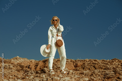 Elegant fashionable woman wearing white linen suit, sunglasses, with round wicker bag, posing on rocks, against the blue sky. Outdoor summer fashion full-length  portrait. Copy, empty space for text