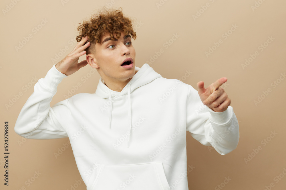 a funny, shocked man stands on a beige background in a white hoodie and with his mouth wide open points his fingers towards an empty space in the background a funny, shocked man stands on a beige background in a white hoodie and with his mouth wide open points his fingers towards an empty space in the background