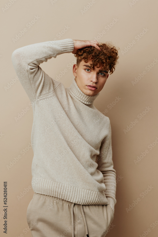 a close portrait of a handsome man with curly hair, standing on a beige background in a light turtleneck and looking at the camera with his hands in his pants pockets, while the other touches his hair a close portrait of a handsome man with curly hair, standing on a beige background in a light turtleneck and looking at the camera with his hands in his pants pockets, while the other touches his hair