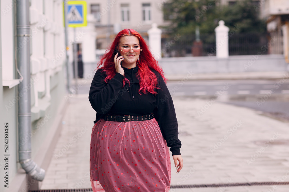 European plus size woman use mobile phone outdoor. Young red pink haired body positive girl walk at city street outdoors