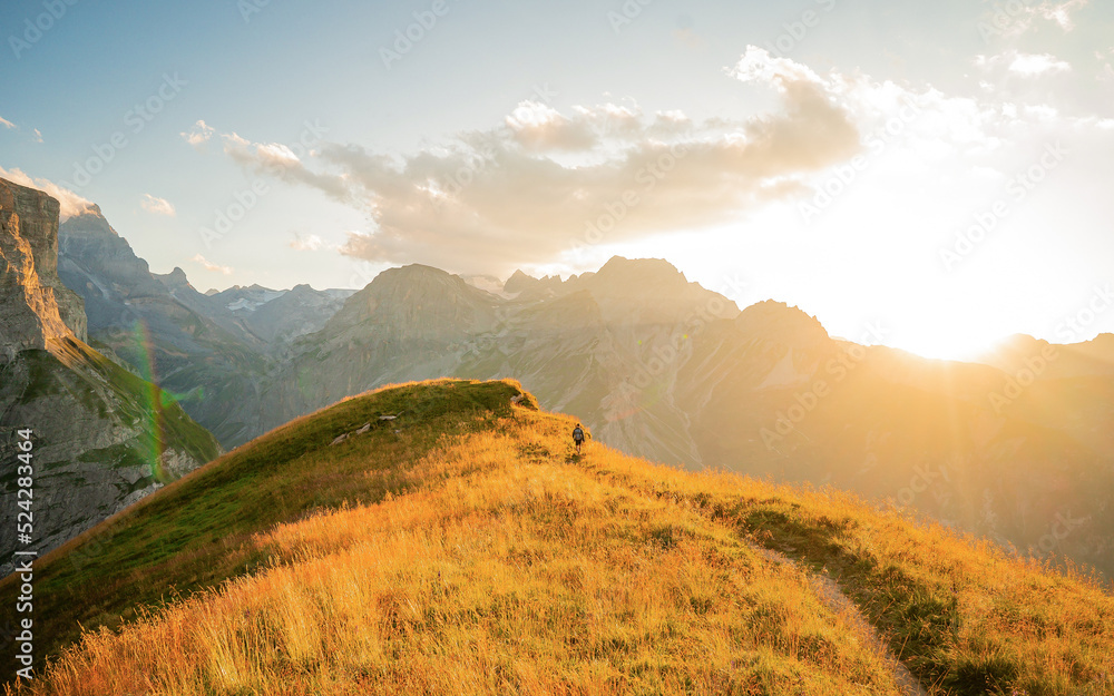 A lone hiker walking through the impressive mountain landscape of the Alps in summer with the sun rising over the mountain peaks in the distance.