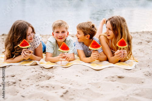 Group of four different age children eating watermelon lying on beach. Two girls and two boys friends holding slice of watermelon on stick enjoy summer together. Friendship, good mood, leisure. photo
