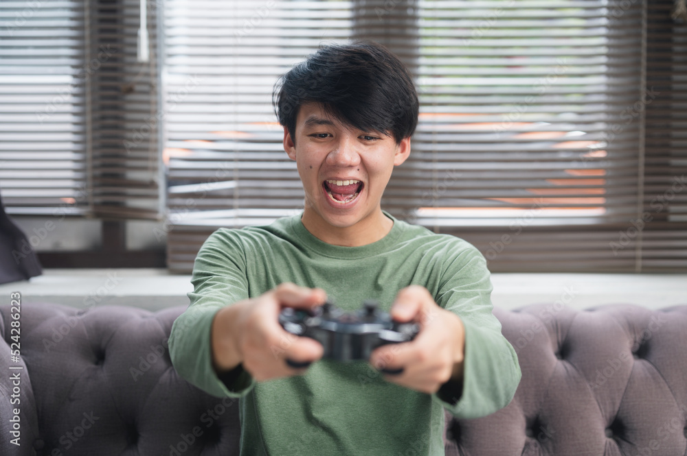 Excited face of Asian gamer or winner gamer holding joystick playing video game online sitting on sofa at living room.