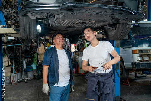 Senior mechanic and young man help to fix the problem of car in area under the car in garage.