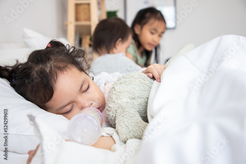 Adorable little child is sleeping in the bed with sister and her toy. The girl is hugging the teddy bear with bottle of milk.