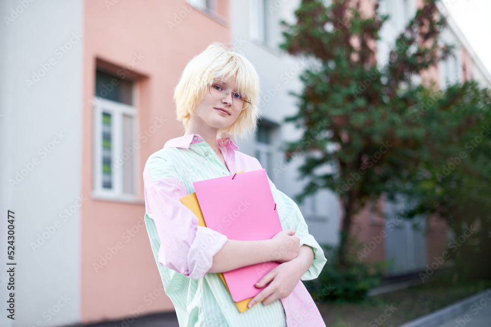 girl schoolgirl in glasses and stylish colored striped shirt holding folders with notebooks in the background of school facade girl schoolgirl in glasses and stylish colored striped shirt holding folders with notebooks in the background of school facade