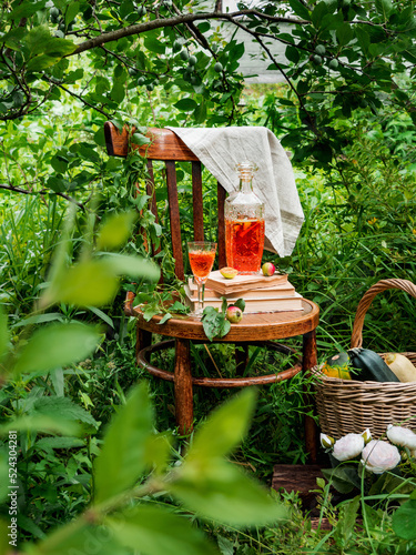 Outdoor picnic in the forest. There is a decanter with orange compote on the books on the chair. Around foliage, greenery. Lazy photo. Wicker basket with a crop of zucchini. Flowers. Rustic style in n