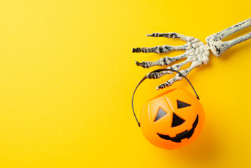 Halloween concept. Top view photo of skeleton hand holding pumpkin basket for trick-or-treat on isolated yellow background with copyspace