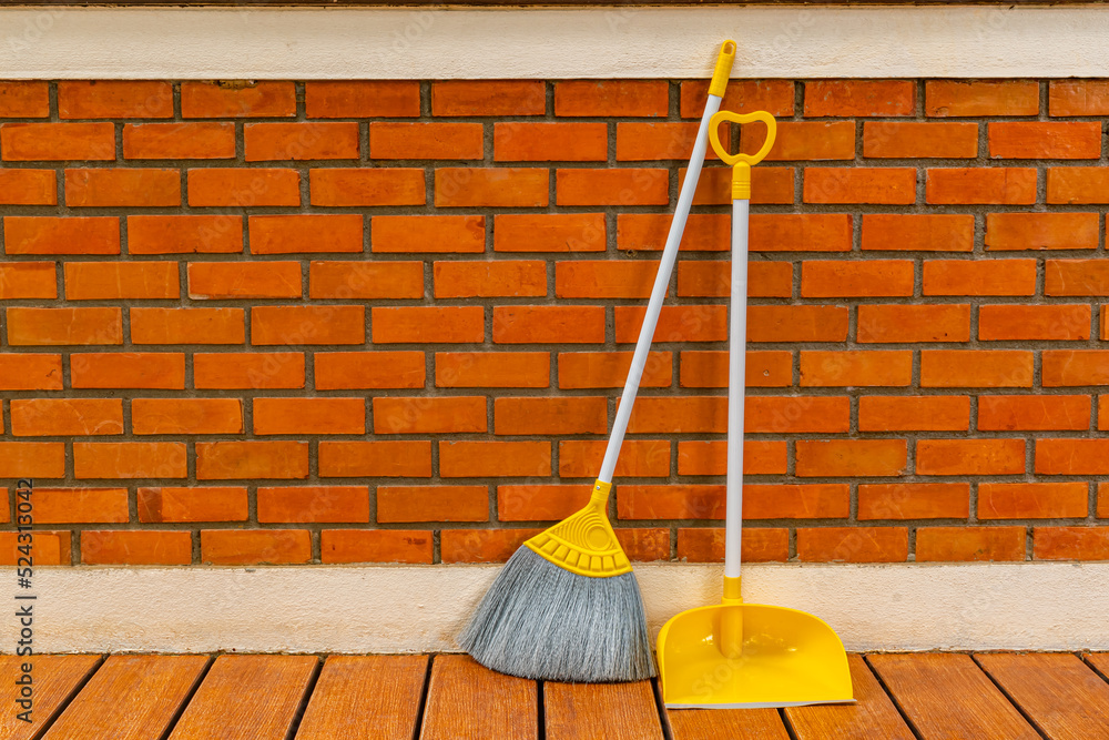 Long handle plastic yellow broom and dustpan prop against a red brick wall of a house. - obrazy, fototapety, plakaty 