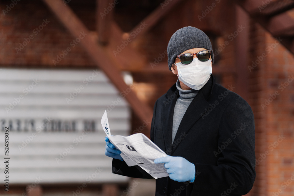 Horizontal shot of man uses virus preventive measures during coronavirus pandemic, wears face medical mask and rubber gloves, reads newspaper. Epidemic disease, Covid-19, health care concept
