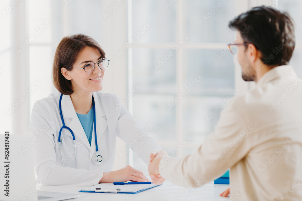 Friendly female doctor greets patient with handshake, pose in private clinic, sit opposite each other, have talk, agree to sign insurance contract, meet in hospital. Health care and medicine concept Friendly female doctor greets patient with handshake, pose in private clinic, sit opposite each other, have talk, agree to sign insurance contract, meet in hospital. Health care and medicine concept