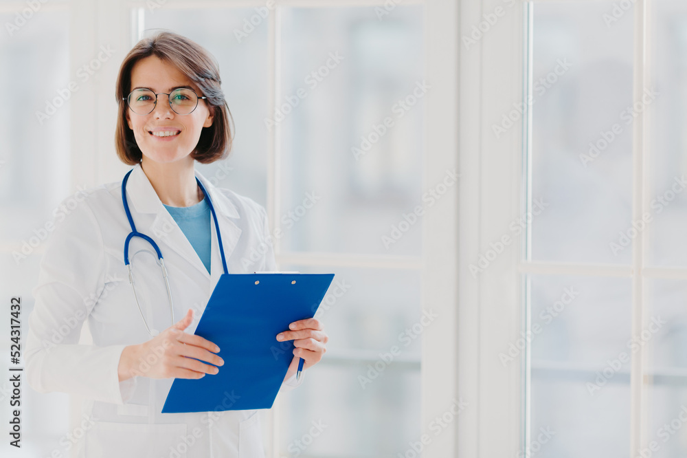Smiling female doctor stands with clipboard, makes prescription for patinet, wears white gown, poses indoor against big window, works in hospital, holds medical record. Copy space for your information Smiling female doctor stands with clipboard, makes prescription for patinet, wears white gown, poses indoor against big window, works in hospital, holds medical record. Copy space for your information