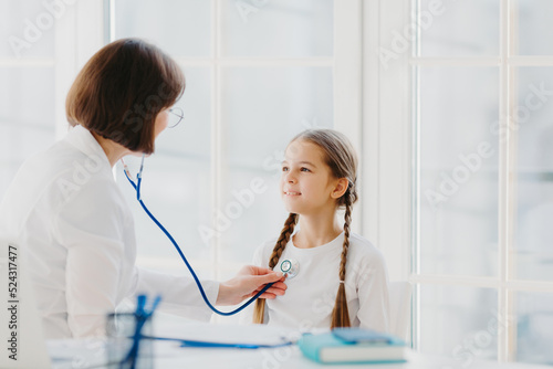 Lovely small girl listens consult of professional experienced doctor who listens her lungs with stethoscope  comes on medical checkup appointment. Children healthcare and clinic visit concept