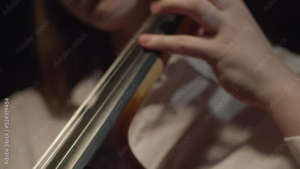 Young female classical musician in a white shirt playing music on a cello. Closeup of a hand on a string instrument, fingers stopping strings, indoors orchestra concert rehearsal Young female classical musician in a white shirt playing music on a cello. Closeup of a hand on a string instrument, fingers stopping strings, indoors orchestra concert rehearsal