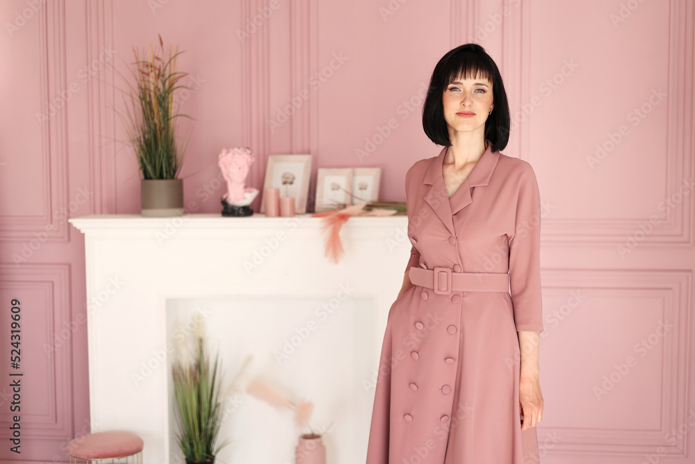 Portrait of attractive smiling brunette with short hair woman folded arms staying alone at stylish interior indoors. Female in soft pink dress is looking in camera Portrait of attractive smiling brunette with short hair woman folded arms staying alone at stylish interior indoors. Female in soft pink dress is looking in camera