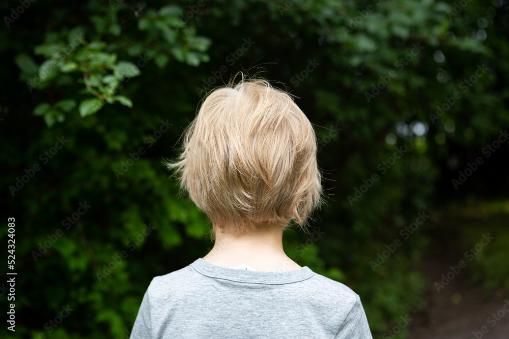 The child's upper body is photographed from behind, with dark green leafy trees in the background. The child's appearance is unisex. The child's upper body is photographed from behind, with dark green leafy trees in the background. The child's appearance is unisex.