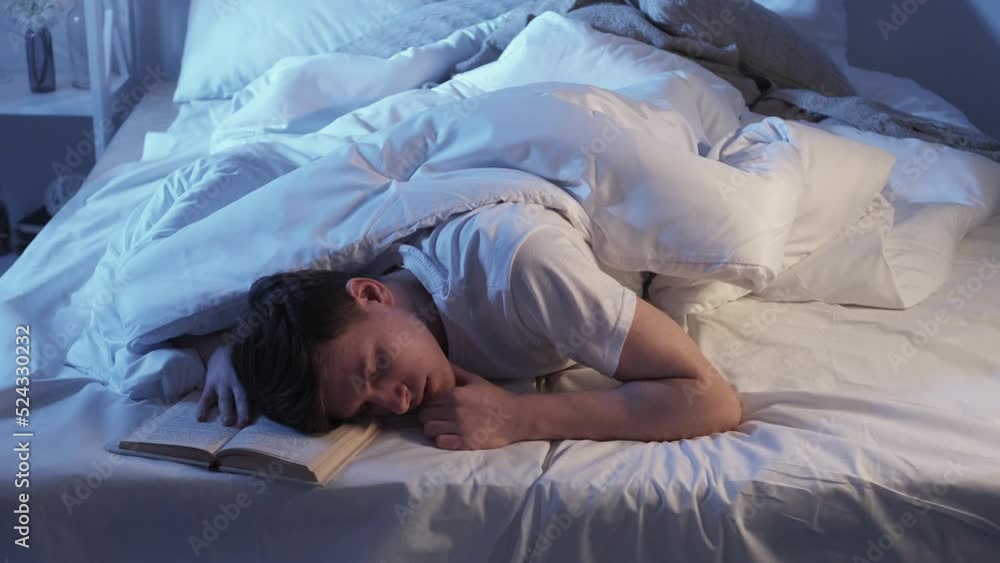 Night relaxing reading. Bedtime habit. Late home study. Tired exhausted man sleeping with open book under blanket in bed in dark bedroom.