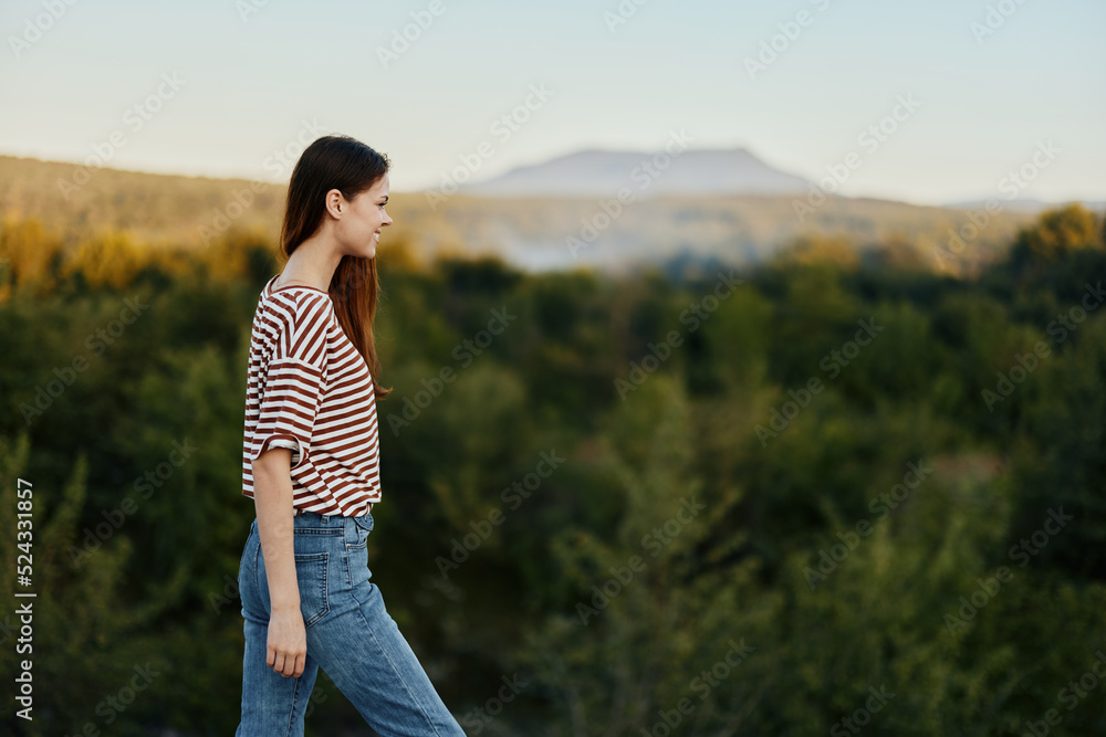 A woman in nature stands with her back to the camera near a cliff and looks at beautiful mountains and trees in the sunset light. travel lifestyle