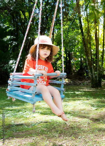 Criança, menina, com roupa laranjada, em um balanço de cordas em meio à natureza. photo