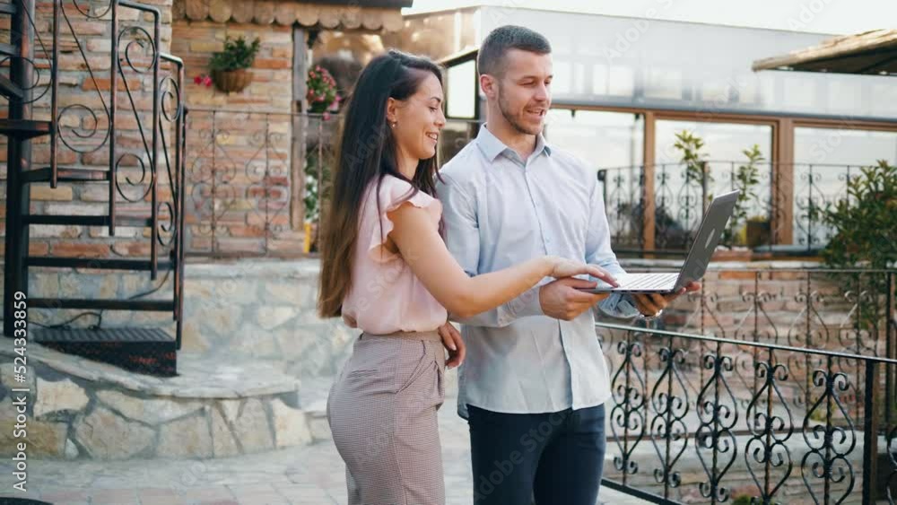 Male real estate working with a laptop computer while standing yard of a modern house and talking to the female customer Male real estate working with a laptop computer while standing yard of a modern house and talking to the female customer
