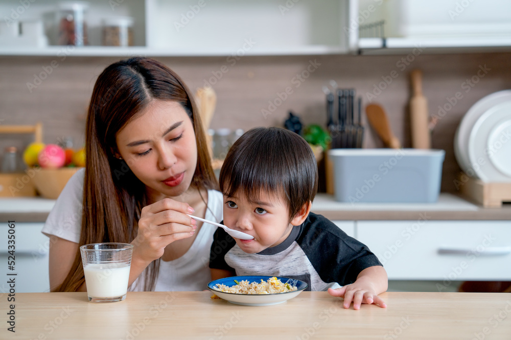Asian mother help her son to eat food with enter and support with her hands and stay together in kitchen of their house and also prepare a glass of milk on table. Asian mother help her son to eat food with enter and support with her hands and stay together in kitchen of their house and also prepare a glass of milk on table.