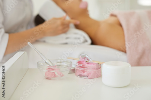 Skin care products at beauty salon. Closeup shot of white plastic jar with luxury face cream and glass bowl with pink kaolin clay or strawberry facial mask placed on table at modern spa center