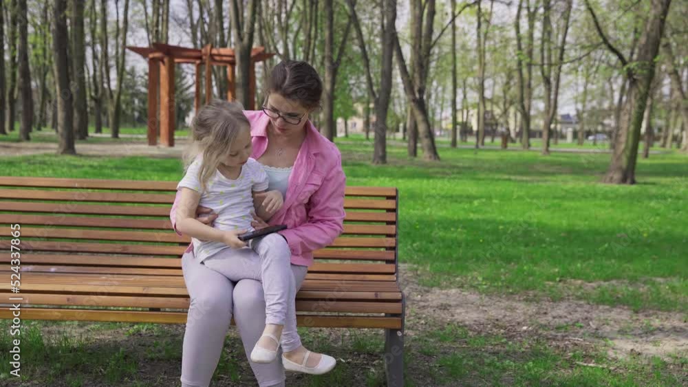 A mother and child sit on a bench and both look at a smartphone. A mother and child sit on a bench and both look at a smartphone.