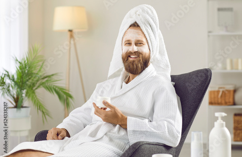 Happy, cheerful, smiling handsome unshaven ginger man in fresh clean white bathrobe and towel enjoying spa and self care day at home and relaxing in comfy chair with kaolin clay spa mask on his face photo