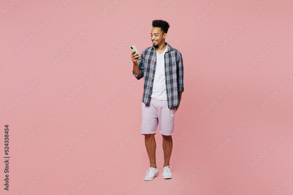 Full body young smiling man of African American ethnicity he wear blue shirt hold in hand use mobile cell phone isolated on plain pastel light pink background studio portrait People lifestyle concept Full body young smiling man of African American ethnicity he wear blue shirt hold in hand use mobile cell phone isolated on plain pastel light pink background studio portrait People lifestyle concept