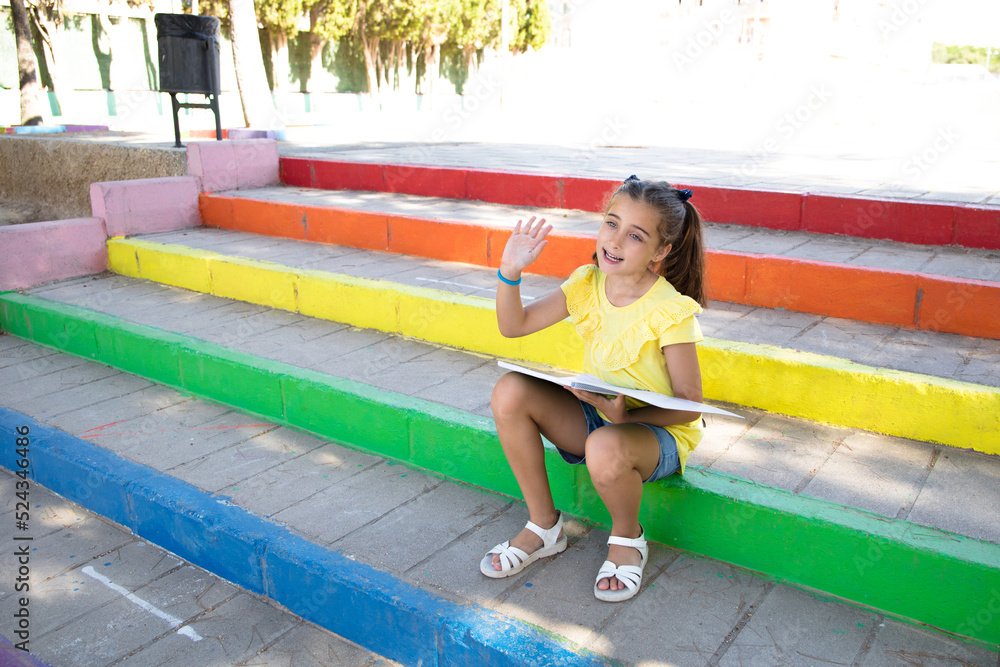 Fototapeta premium Cute girl sitting on a staircase with the colors of the LGTBI flag, picking up a notebook.